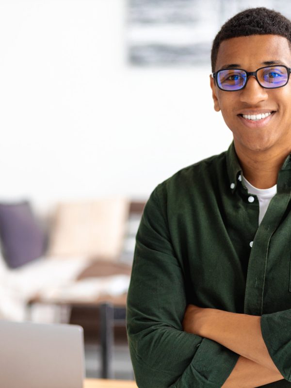 Smiling successful businessman with eyeglasses stands with crossed arms smiling and looking at the camera. Young male confident entrepreneur or small business owner in office, copy space