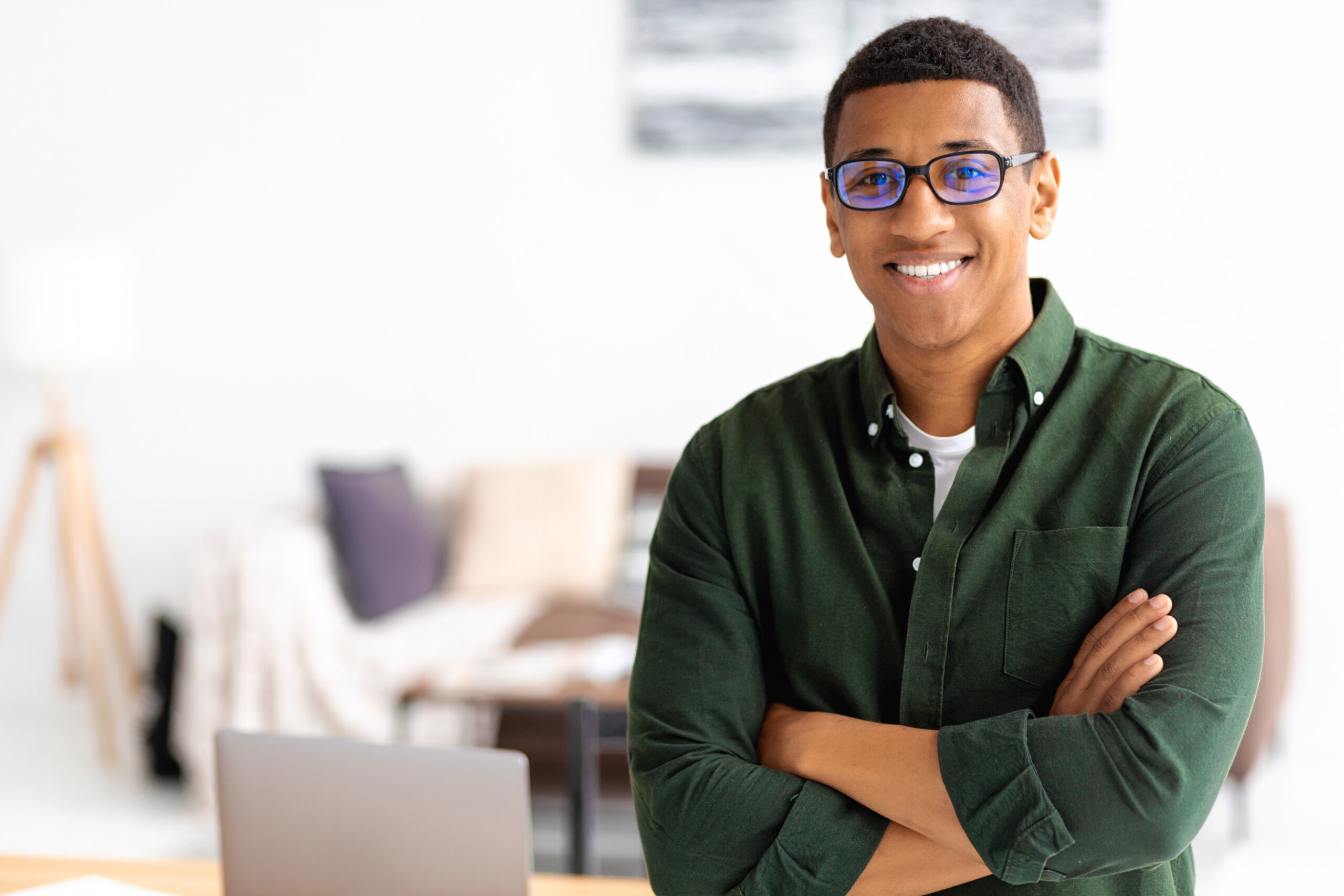 Smiling successful businessman with eyeglasses stands with crossed arms smiling and looking at the camera. Young male confident entrepreneur or small business owner in office, copy space